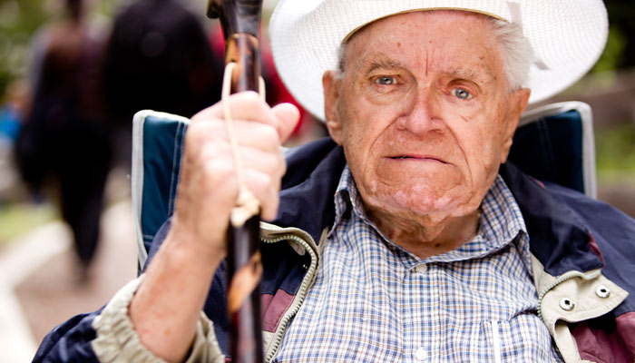 An elderly man wearing a white hat and plaid shirt looks determined as he holds onto a walking stick, sitting outdoors in a blue chair.