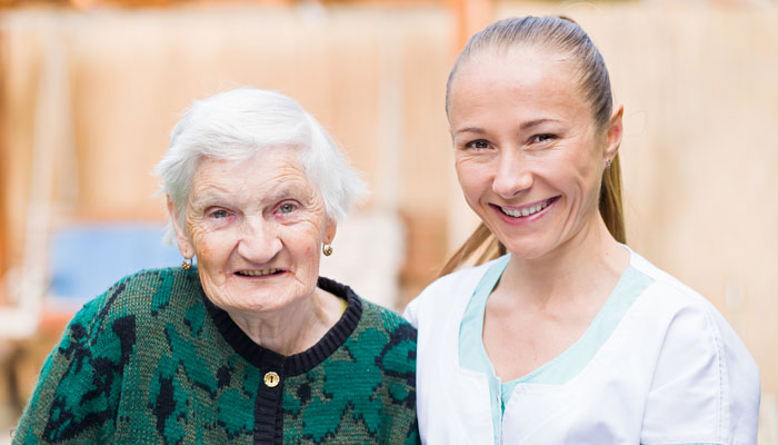 An elderly woman with white hair smiles broadly next to a younger woman in a nurse's uniform, both posing for a photo in a warmly lit setting.