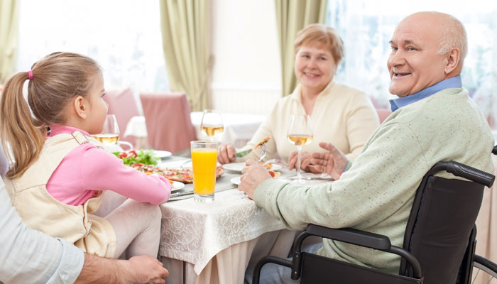 A joyful family meal with a young girl, a woman, and an elderly man in a wheelchair, sitting at a dining table with glasses of juice and plates of food, smiling and engaging in conversation.