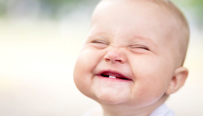 A joyful baby with a wide, toothless smile and closed eyes, basking in a softly blurred sunny background.