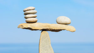 A balanced stone sculpture featuring smooth rocks stacked on top of a flat rock balanced on a single upright stone, set against a clear blue sky.
