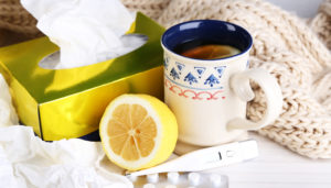 A cozy scene with a steaming mug of tea, a halved lemon, a thermometer, and a box of tissues surrounded by a soft, knitted scarf, suggesting cold or flu treatment.