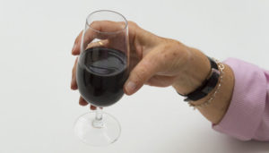 A hand holding a glass of red wine, with a focus on the glass and the person's wrist wearing a silver bracelet. the background is plain and light-colored.