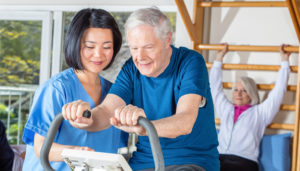 An elderly man exercises on a stationary bike with assistance from a female nurse in a rehab center, while another elderly woman exercises in the background.