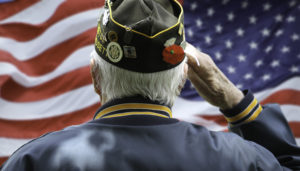An elderly veteran in a decorated service cap salutes, with an american flag in the background.