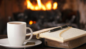 A cozy scene featuring a white cup of coffee next to an open notebook with handwritten notes and a pen, with a warm fireplace in the background.