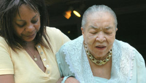 An elderly woman in a light blue blouse, assisted by a younger woman in a yellow top, walks carefully down stairs with eyes closed and a pained expression.