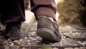 Close-up of a person walking on a pebble path, focusing on worn-out shoes with frayed edges and soles visible from behind. one foot is stepping forward, mid-stride.