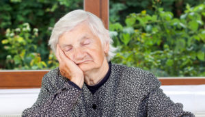 An elderly woman with closed eyes, resting her cheek on her hand, sits by a window overlooking a lush green garden. she wears a patterned blouse.