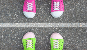 Two pairs of colorful sneakers facing each other on asphalt, one pair pink and the other green, with a white line between them.
