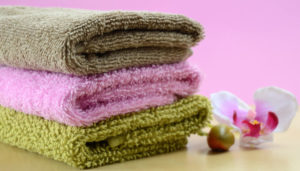 Three neatly stacked towels in green, pink, and beige colors with an orchid flower and a small round seed beside them on a wooden surface against a pink background.