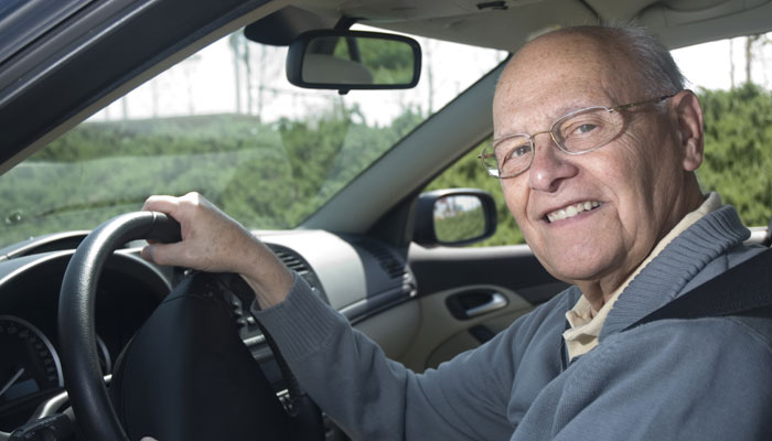 An elderly man smiling while driving a car, his hands on the steering wheel, with light entering through the car's window.