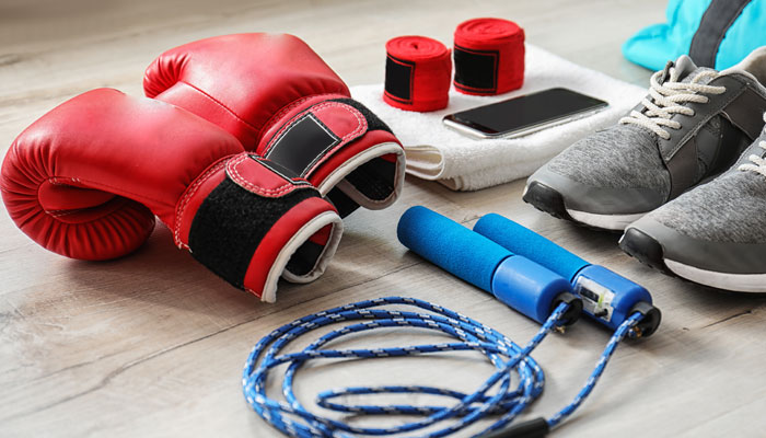 Assorted gym equipment on a wooden floor, including red boxing gloves, hand wraps, a jump rope, trainers, a towel, and a smartphone.