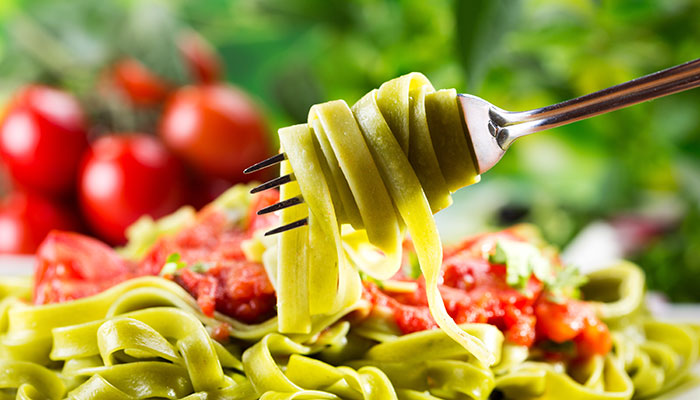 A fork twirls a serving of green fettuccine pasta with chunks of tomato and herbs, set against a backdrop of fresh tomatoes.