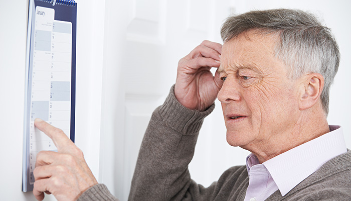 An elderly man looks puzzled while pointing at a wall-mounted calendar and scratching his head in an indoor setting.
