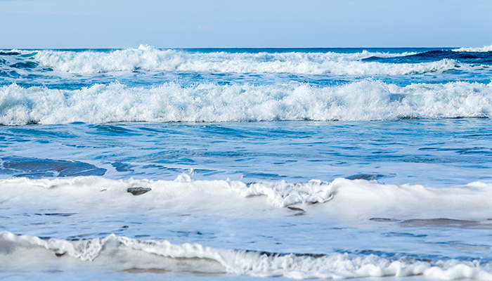 Vibrant ocean waves rolling onto a shore, with white foam and clear blue sky.