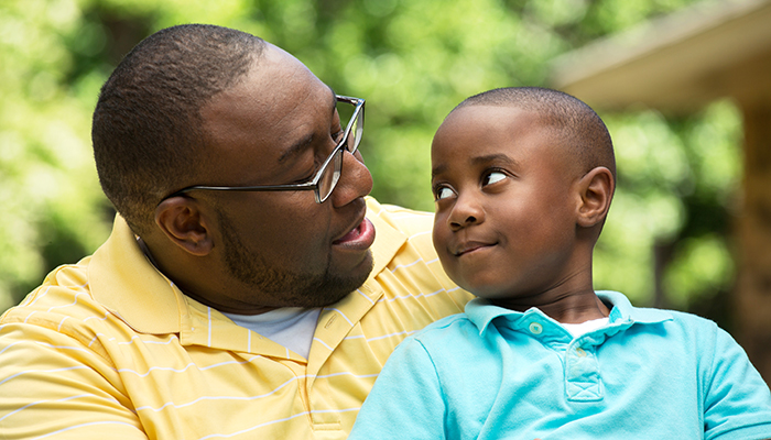 A father and his young son, both smiling, share a loving gaze in a park setting, surrounded by lush greenery. they are dressed in casual, colorful clothing.