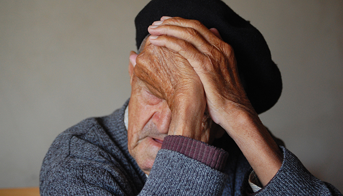 An elderly man wearing a beret and a sweater, with his face partially covered by his hands, appears in deep thought or distress.