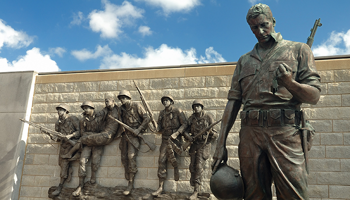 Bronze sculpture of a group of soldiers, one in the foreground holding a helmet, with others behind holding guns against a cloudy sky backdrop.