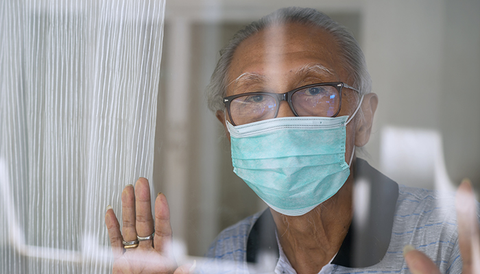 Elderly man wearing a surgical mask looking through a window with a hopeful expression, hands gently touching the glass.