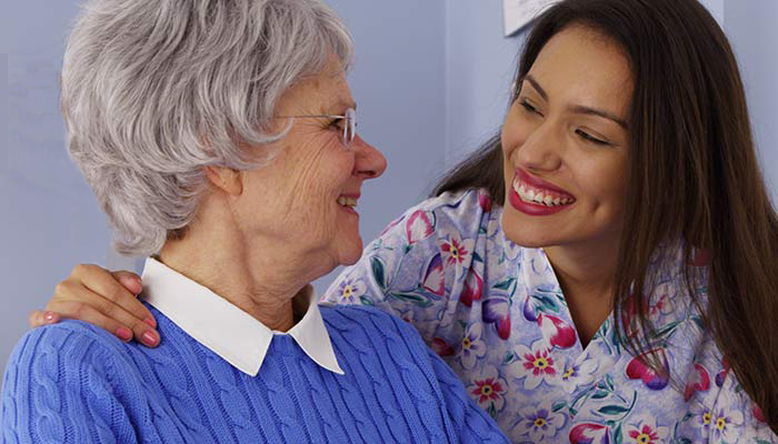 An elderly white-haired woman in a blue sweater and a younger woman with long brown hair in a floral shirt smile at each other, sharing a joyful moment.