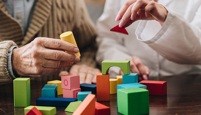 Two individuals, one elderly and one younger, sit at a table playing with colorful wooden blocks, carefully placing them to create structures.