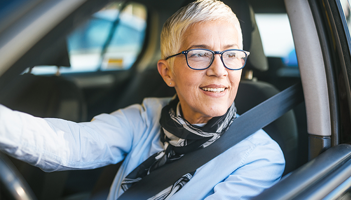 A joyful senior woman with short white hair and glasses, wearing a scarf and a seatbelt, smiling while driving a car.