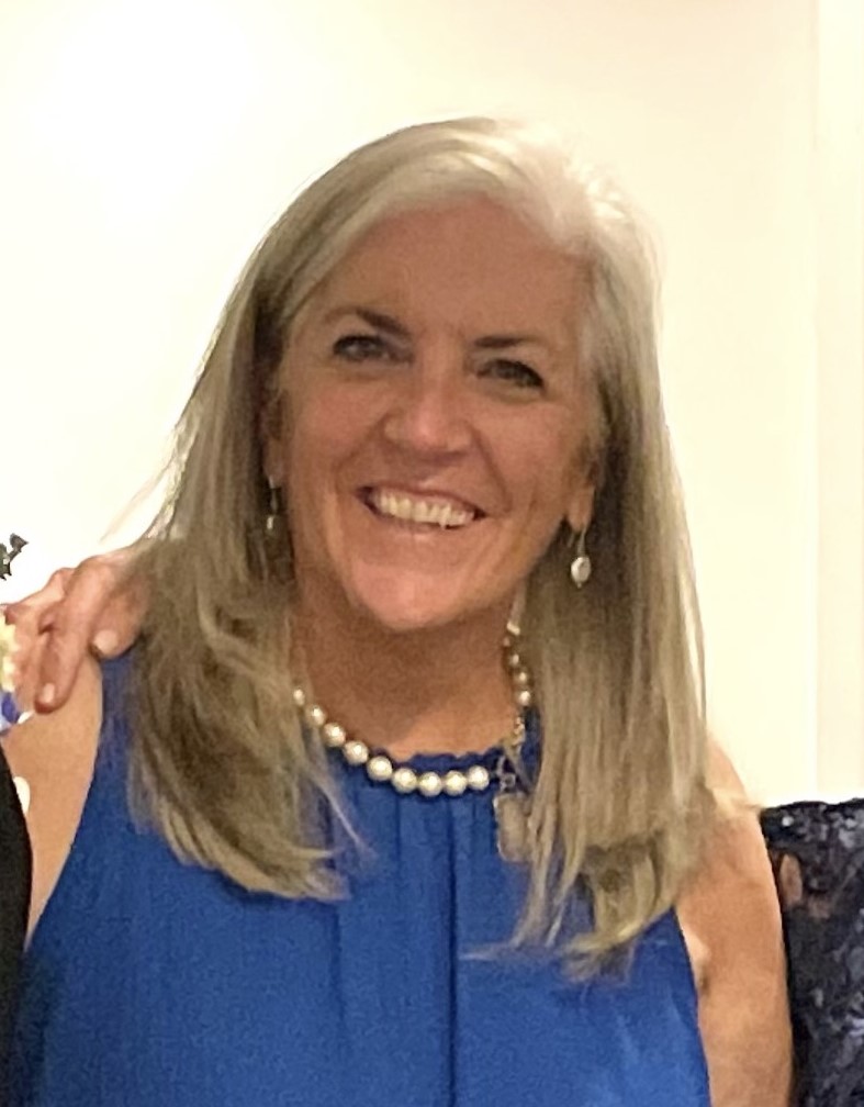 A woman with long, gray hair smiles at the camera. She is wearing a sleeveless blue top, a pearl necklace, matching earrings, and is standing in front of a neutral background.